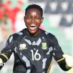 Andile Dlamini wears a black South African goalkeeping jersey with grey trim along with white gloves, as she clenches her fists and opens her mouth wide to scream towards the camera