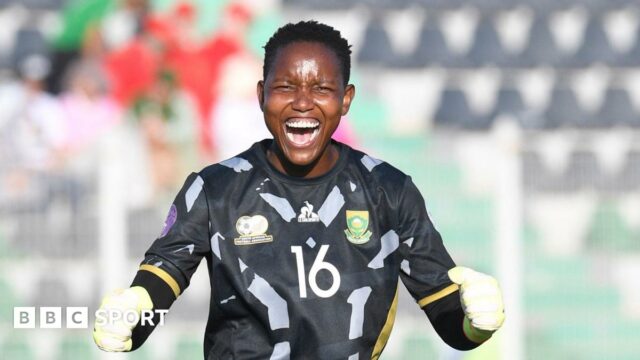 Andile Dlamini wears a black South African goalkeeping jersey with grey trim along with white gloves, as she clenches her fists and opens her mouth wide to scream towards the camera