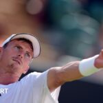 Billy Harris, who is wearing a white T-shirt, cap and wristband, throwing a ball into the air to serve. His left arm is outstretched and he has a look of concentration on his face as he focuses on the ball.
