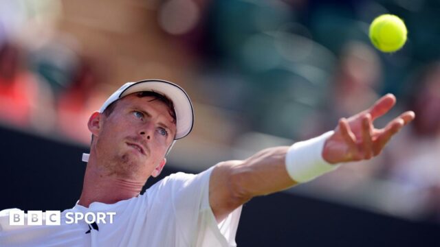 Billy Harris, who is wearing a white T-shirt, cap and wristband, throwing a ball into the air to serve. His left arm is outstretched and he has a look of concentration on his face as he focuses on the ball.