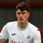 Joel Cotterill standing with the ball at his feet and his right arm held out during a pre-season match for Swansea