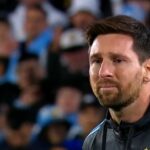 Lionel Messi v drese Argentíny sa posúva na futbalovom poli a jeho nohou ovláda loptu. Hráč v gaštanovej Venezuele Jersey k nemu beží. Rozhodca v žltej uniforme stojí v blízkosti trávy.