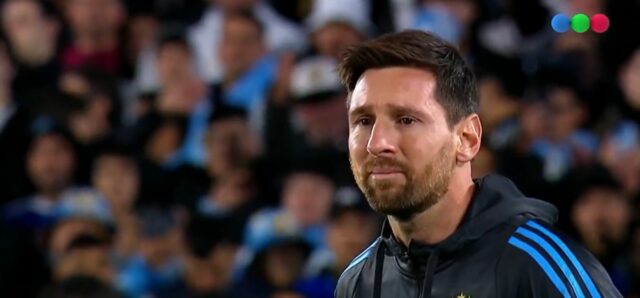Lionel Messi v drese Argentíny sa posúva na futbalovom poli a jeho nohou ovláda loptu. Hráč v gaštanovej Venezuele Jersey k nemu beží. Rozhodca v žltej uniforme stojí v blízkosti trávy.