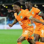 Futbalista Amad Diallo v oranžovom drese so zeleným lemom a bielym číslom 15 vpredu a oranžovými šortkami roztiahne ruky nabok a beží oslavovať po skórovaní Pobrežia Slonoviny proti Kamerunu na Afcon 2025. Za Amadom je vidieť tímového kolegu Vakouna Baya, škerí sa a ukazuje pravou rukou natiahnutou vpredu. Obaja hráči majú obuté ružové čižmy s modrými pruhmi. Bielu bránku je možné vidieť rozmazane v pozadí na sviežom zelenom ihrisku, zatiaľ čo rozmazané svetlá je možné vidieť aj v tmavom dave.