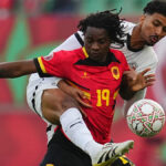 Futbalista Fredy v červenom tričku so žltými detailmi a jeho menom a číslom 16 na chrbte v žltej farbe strieľa loptu z priameho kopu pre Angolu proti Egyptu na Afcon 2025. Piati hráči Egypta v bielom drese s čiernym lemovaním a čiernymi číslami vpredu vyskočili do vzduchu a pokúsili sa zachytiť loptu, zatiaľ čo Fredyho spoluhráč na chrbte, Maestro, sa pozerá na strelu.