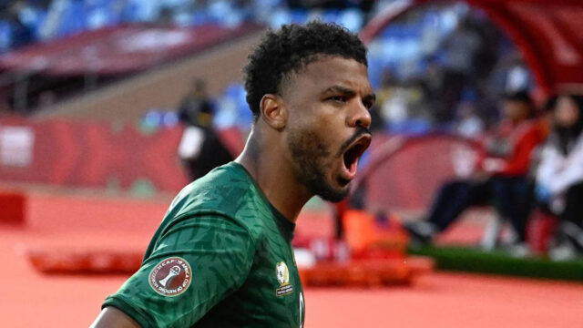 Usmiaty Oswin Appollis, ktorý je viditeľný od pása nahor, má na sebe zelený dres s krátkym rukávom z Juhoafrickej republiky, zdobený zlatou a s bielym číslom sedem v strede hrude, beží a rozpaží ruky nízko vedľa seba, aby oslávil gól proti Zimbabwe na Afcon 2025. Za ním a naľavo od obrazu je vidieť dvoch spoluhráčov z Južnej Afriky, ktorí ho bežia, aby ho chytili