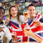 Muži a ženy pijú pivo a oslavujú úspech športového tímu, mávajú vlajkou Veľkej Británie, muž ukazuje víťazný znak v bare