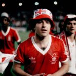 Skupina jednotlivcov oblečených v červených futbalových uniformách stojí na ihrisku, pričom niektorí z nich majú čiapky. Jednotlivec v strede sa pozerá doľava, zatiaľ čo ostatní v pozadí sa zapájajú do rozhovoru. Zdá sa, že ide o športovú udalosť alebo tréning.