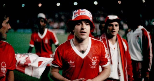 Skupina jednotlivcov oblečených v červených futbalových uniformách stojí na ihrisku, pričom niektorí z nich majú čiapky. Jednotlivec v strede sa pozerá doľava, zatiaľ čo ostatní v pozadí sa zapájajú do rozhovoru. Zdá sa, že ide o športovú udalosť alebo tréning.