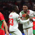 Lassine Sinayoko v bielom drese Mali je vidieť uprostred akcie, keď strieľa penaltu na Africkom pohári národov proti Maroku. Sinayoko práve kopol do lopty, ktorá letí pozdĺž stredu obrazu, len mierne vyvýšená nad sviežim zeleným trávnikom. Päť hráčov je v pozadí na hranici pokutového územia, vrátane dvoch hráčov Mali, z ktorých jeden má ruky zopnuté v modlitbovom geste. Dvaja z troch marockých hráčov v červeno-zelenom už vtrhnú do lóže v očakávaní