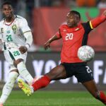 Futbalista Christian Kofane v bielom drese so zelenými, žltými a červenými detailmi a s číslom 26 v zelenom na hrudi a stehne strieľa gól za kamerunské národné mužstvo proti Mozambiku na Afcon 2025. Mozambický hráč Oliver Keman v červenom tričku s čiernymi detailmi a čiernymi šortkami, obaja s číslom 20 na nich v bielej farbe, sa rúti cez nohu cez nohu Kofanecept. loptu, ale je dobre vidieť za ním