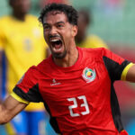 Diogo Calila zobrazený od pása nahor sa otáča na oslave po strelení gólu Mozambiku proti Gabonu na Afcon 2025. Má na sebe červený dres s krátkym rukávom s čierno-žltým lemom a bielym číslom 23 v strede hrude. Calila má tenkú briadku bradu a fúzy a dokorán otvára ústa, aby kričal od radosti, pričom rozťahuje obe ruky nabok a zatína päste. Hráči Gabonu v žltej a modrej súprave sú rozmazaní v pozadí