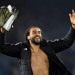 Jednotlivec, čiastočne bez košele, dvíha ruky, zatiaľ čo drží trofej, stojí na otvorenom poli s rozmazaným pozadím, pravdepodobne počas slávnostnej udalosti.