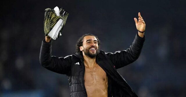 Jednotlivec, čiastočne bez košele, dvíha ruky, zatiaľ čo drží trofej, stojí na otvorenom poli s rozmazaným pozadím, pravdepodobne počas slávnostnej udalosti.