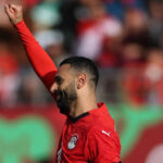 Mohamed Salah v červenom egyptskom drese, bielych trenírkach a čiernych štulpniach strieľa ľavou nohou a strieľa pokutový kop proti Juhoafrickej republike na Afcon 2025. Lopta je vidieť nízko na obrázku úplne vpravo a smeruje k bráne, ktorú nie je vidieť. Mierne rozmazané pozadie sú dvaja hráči Egypta a traja hráči Juhoafrickej republiky v zelenom a žltom, ktorí sa práve začínajú presúvať do pokutového územia po tom, čo čakali na okraji 18-yardového poľa.