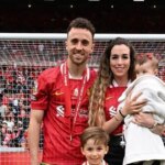 Mužský športovec v červenom drese triumfálne stojí so zdvihnutými rukami a zdanlivo oslavuje významný úspech na ihrisku. Pozadie je plné nadšeného davu, čo naznačuje dôležitosť okamihu.