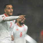 Futbalista Elias Achouri oslavuje po strelení gólu v zápase medzi Tuniskom a Ugandou na Afcon 2025. V bielom drese je viditeľný od hrude nahor a naťahuje pravú ruku dopredu, aby ukázal ukazovákom. Tímového kolegu je vidieť od krku nahor rozmazané v pozadí a je vidieť padať veľké kvapky dažďa