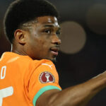 Amad Diallo, pri pohľade od hrude nahor, oblečený v oranžovej košeli Pobrežia Slonoviny so zeleným lemom, vyčarí na tvári mierny úsmev. V rozmazanom pozadí za ním je vidieť časť bránky, sieť a chrbát hráča Burkiny Faso v bielom