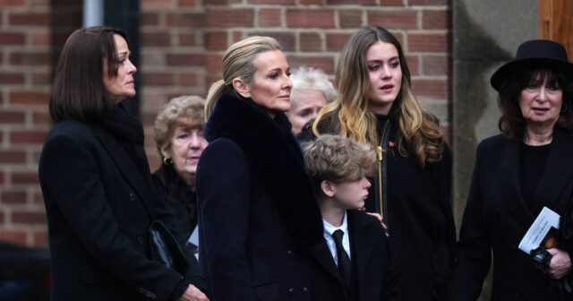 Dcéra Gabby Logan necháva návštevníkov pohrebu v slzách, keď plní Gabby Logan a jej rodina v formálnom oblečení stoja pred tehlovou stenou na pohrebe Terryho Yoratha.