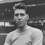 Čiernobiela snímka zobrazujúca anglického brankára Colina McDonalda, ako vyskakuje do vzduchu so zdvihnutou pravou rukou, keď bojuje o loptu počas priateľského zápasu na štadióne Wembley v októbri 1958. Na sebe má svetlý jednoliaty dres, tmavé šortky a ponožky, ako aj pár tmavých rukavíc. Za loptou skáče aj číslo 5 Anglicka. V pozadí na tribúnach štadióna vidno tisíce ľudí. Z jednej z pozemných veží visí vlajka odborového zväzu.