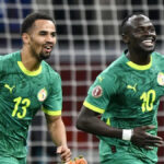 Sadio Mane oslavuje s úsmevom na tvári, keď ho prenasleduje tímový kolega zo Senegalu Iliman Ndiaye. Obaja hráči majú na sebe zelené senegalské dresy, zatiaľ čo egyptský brankár Mohamed El Shenawy vo fialovom drese je skľúčený v pozadí stojac pred rozmazanou bielou sieťou.