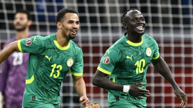 Sadio Mane oslavuje s úsmevom na tvári, keď ho prenasleduje tímový kolega zo Senegalu Iliman Ndiaye. Obaja hráči majú na sebe zelené senegalské dresy, zatiaľ čo egyptský brankár Mohamed El Shenawy vo fialovom drese je skľúčený v pozadí stojac pred rozmazanou bielou sieťou.