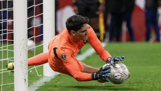 Marocký brankár Yassine Bounou chytá penaltu nízko po svojej pravej strane. V oranžovej súprave a čierno-modrých rukaviciach je vidieť, ako mu rozmazaná lopta udiera do rúk, keď sa nachádza tesne nad trávnikom na svojej bránkovej čiare. V pozadí je vidieť nigérijského brankára Stanleyho Nwabaliho v žltom, ako kľačí dopredu s hlavou na trávniku