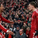 Dvaja futbalisti v červených uniformách oslavujú gól pred nadšeným publikom, pričom niekoľko fanúšikov v pozadí má na sebe červené šatky.