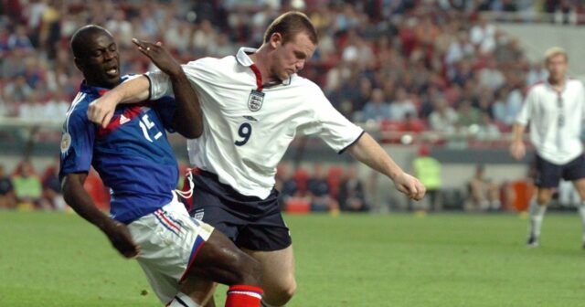 Dvaja futbalisti sú zapojení do dynamického a intenzívneho zápasu na ihrisku. Jeden hráč v modrom drese urputne zápasí s druhým v bielom drese. Obaja sú pripravení zasiahnuť loptu, ktorá je vo vzduchu, zatiaľ čo ostatní hráči a diváci sú viditeľní v pozadí.