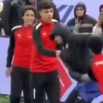 Skupina jednotlivcov oblečených v červeno-čiernych uniformách vystupuje na ihrisku s veľkým transparentom zobrazujúcim slovo