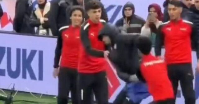 Skupina jednotlivcov oblečených v červeno-čiernych uniformách vystupuje na ihrisku s veľkým transparentom zobrazujúcim slovo 