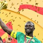 Sadio Mane oslavuje s úsmevom na tvári, keď ho prenasleduje tímový kolega zo Senegalu Iliman Ndiaye. Obaja hráči majú na sebe zelené senegalské dresy, zatiaľ čo egyptský brankár Mohamed El Shenawy vo fialovom drese je skľúčený v pozadí stojac pred bielou sieťou.