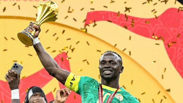 Sadio Mane oslavuje s úsmevom na tvári, keď ho prenasleduje tímový kolega zo Senegalu Iliman Ndiaye. Obaja hráči majú na sebe zelené senegalské dresy, zatiaľ čo egyptský brankár Mohamed El Shenawy vo fialovom drese je skľúčený v pozadí stojac pred bielou sieťou.