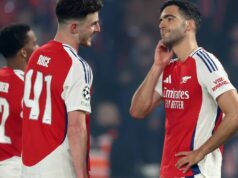 Hviezda Arsenalu Mikel Merino zdieľa príspevok na nemocničnom lôžku, zatiaľ čo Declan Rice posiela podpornú správu Traja futbalisti oblečení v červeno-bielych dresoch stoja na ihrisku zabraní do rozhovoru s rozostreným pozadím divákov.