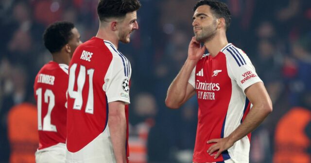 Traja futbalisti oblečení v červeno-bielych dresoch stoja na ihrisku zabraní do rozhovoru s rozostreným pozadím divákov.