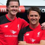 Dve osoby oblečené v červených dresoch stoja blízko pri sebe a vyzerajú ako v oslavnej póze s transparentom v pozadí s nápisom