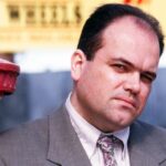 Barry z EastEnders oblečený vo formálnom obleku vrátane kravaty je umiestnený vedľa červeného stĺpa. Na pozadí je banner so žltým textom, hoci konkrétny obsah textu nie je rozpoznateľný.