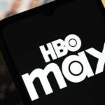 Detailný pohľad na smartfón s logom HBO Max na obrazovke. Pozadie je rozmazané a zobrazuje rôzne zábavné médiá v okolí.