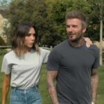 Jednotlivec v sivom tričku a khaki nohaviciach stojí vo vonkajšom prostredí v sprievode osoby v bielom tričku a modrých džínsoch. Na pozadí je dom a svieža zelená krajina.