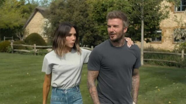 Jednotlivec v sivom tričku a khaki nohaviciach stojí vo vonkajšom prostredí v sprievode osoby v bielom tričku a modrých džínsoch. Na pozadí je dom a svieža zelená krajina.