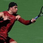 Cameron Norrie v modro-oranžovom tričku oslavuje svoje víťazstvo proti Alexovi de Minaur úsmevom a natiahnutím oboch rúk vedľa seba