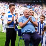 Starší muž v modrej košeli tlieska rukami, keď sedí na štadióne, obklopený davom divákov.