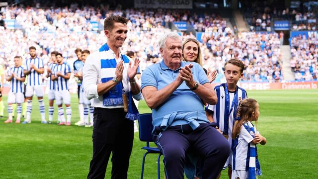 Starší muž v modrej košeli tlieska rukami, keď sedí na štadióne, obklopený davom divákov.