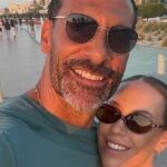 Dvaja jednotlivci stoja spolu na mestskej ulici, obaja majú slnečné okuliare a usmievajú sa. Na pozadí je vydláždená cesta s stĺpikmi, skupina ľudí kráčajúcich v diaľke a rad budov pozdĺž horizontu.