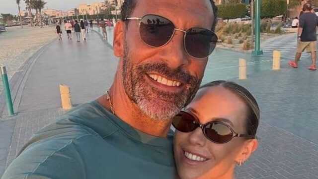 Dvaja jednotlivci stoja spolu na mestskej ulici, obaja majú slnečné okuliare a usmievajú sa. Na pozadí je vydláždená cesta s stĺpikmi, skupina ľudí kráčajúcich v diaľke a rad budov pozdĺž horizontu.