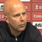 Jednotlivec s holou hlavou v čiernej mikine s kapucňou sedí na tlačovej konferencii a pozorne oslovuje zhromaždené médiá. Za ním kulisa ozdobená rôznymi sponzorskými logami vrátane