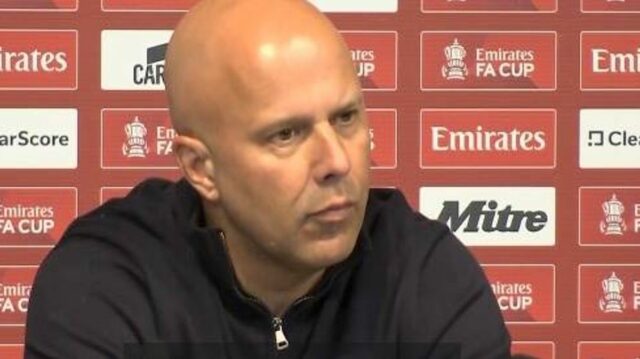 Jednotlivec s holou hlavou v čiernej mikine s kapucňou sedí na tlačovej konferencii a pozorne oslovuje zhromaždené médiá. Za ním kulisa ozdobená rôznymi sponzorskými logami vrátane 