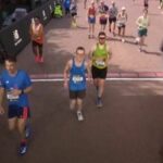 Traja bežci sa zúčastňujú maratónu na vydláždenej mestskej ulici s červenou stenou a reklamami v pozadí. Prítomní sú diváci a bežci sú oblečení v atletickom oblečení so zameraním na preteky.