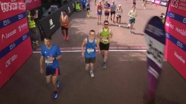 Traja bežci sa zúčastňujú maratónu na vydláždenej mestskej ulici s červenou stenou a reklamami v pozadí. Prítomní sú diváci a bežci sú oblečení v atletickom oblečení so zameraním na preteky.