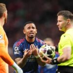 Zdá sa, že dvaja jednotlivci v športových uniformách, z ktorých jeden drží futbalovú loptu, diskutujú s rozhodcom zápasu v rušnom prostredí štadióna s divákmi viditeľnými v pozadí.